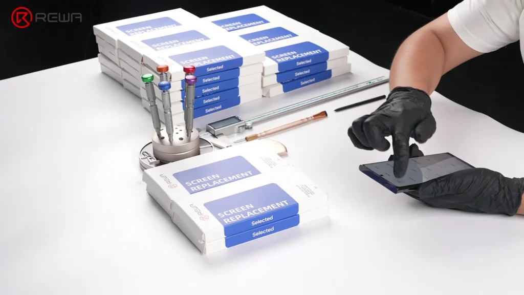 Tecnico che testa lo schermo di uno smartphone accanto a pile di scatole di ricambio per schermi REWA e strumenti di riparazione